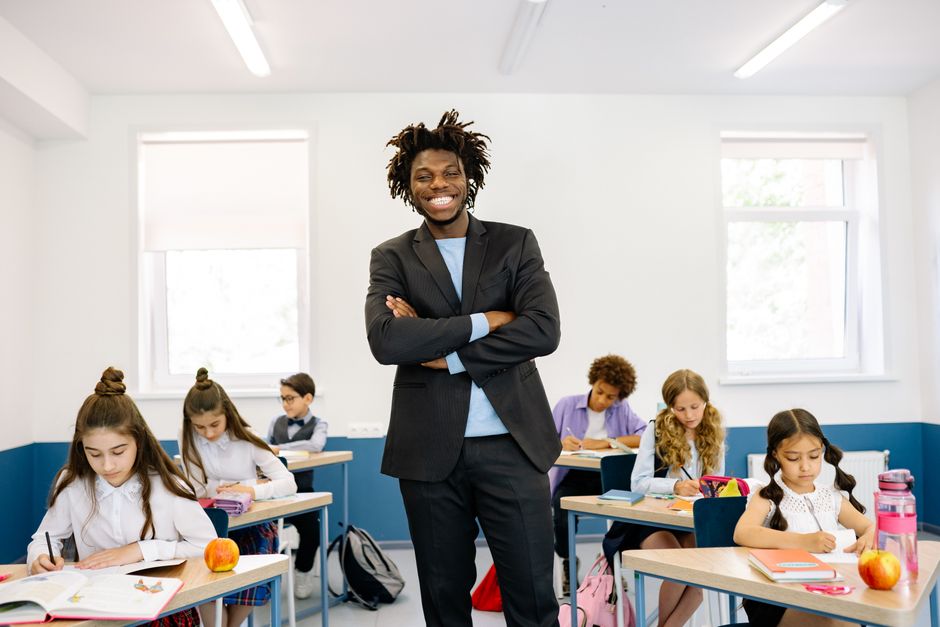 Smiling teacher standing in diverse classroom with students focused on their work.